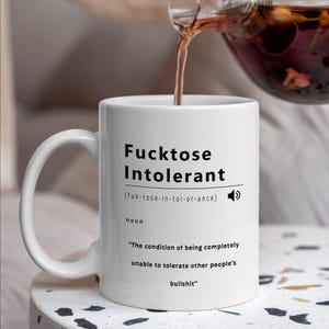 May include: White ceramic mug with the text "Fucktose Intolerant" in black. The mug has a definition of the word and the phrase "The condition of being completely unable to tolerate other people's bullshit". Tea is being poured into the mug.