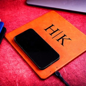 May include: A brown leather charging pad with the initials "H|K" embossed on it. A black smartphone is charging on the pad, and a black charging cable is plugged into the pad.