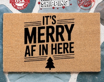 May include: A rectangular doormat with the text "IT'S MERRY AF IN HERE" in black. A black Christmas tree graphic is below the text. The doormat is made of a tan material. The words "MADE IN USA" and "SATISFIED CUSTOMER" are also visible.