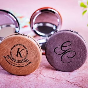 May include: Two compact mirrors with personalized leather cases. One case is brown with the initials "K.K." and the name "KARMEN KATES" engraved. The other case is gray with the initials "G.C." engraved.