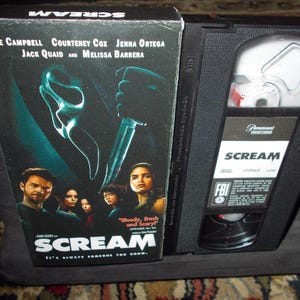 Peut inclure: Une cassette VHS et son boîtier pour le film "Scream" avec Neve Campbell, Courteney Cox, Jenna Ortega, Jack Quaid et Melissa Barrera. La couverture montre un personnage masqué avec un couteau. La cassette est noire.