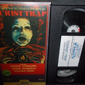 Puede incluir: Una caja de cinta VHS para la película de terror "Tourist Trap". La portada presenta un primer plano de una figura enmascarada con una expresión siniestra. El texto "Every year young people disappear. Tourist Trap" está impreso en letras rojas y negritas. La película está protagonizada por Chuck Connors, Tanya Roberts y Jon Van Ness. La caja es negra con una etiqueta roja y blanca.