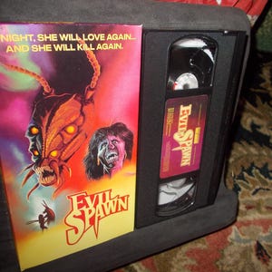 May include: A VHS tape case with a black and red design. The front of the case features a graphic of a monstrous insect with glowing eyes and the text "Evil Spawn". The back of the case has the text "Tonight, she will love again... and she will kill again."