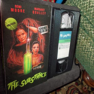 May include: A VHS tape case with a black background and a colourful image of two women. The title of the film is "The Substance" and the actors are Demi Moore and Margaret Qualley. The case also includes the words "Horror" and "Remember You Are One".