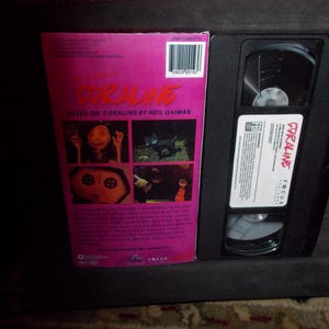 May include: A VHS video cassette and its case for the film "Coraline." The case is pink with images from the movie and the title in red. The VHS tape is black with a white label that reads "Coraline."
