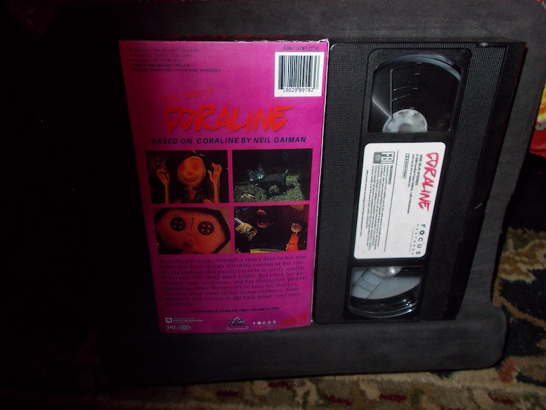 May include: A VHS video cassette and its case for the film "Coraline." The case is pink with images from the movie and the title in red. The VHS tape is black with a white label that reads "Coraline."