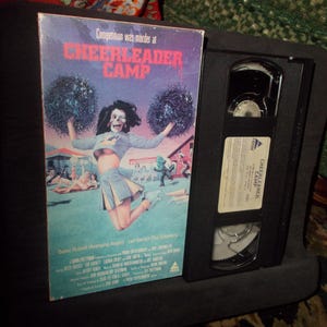 May include: A VHS tape case with a black and white image of a woman in a cheerleader outfit. The text on the case reads "Competition was murder at CHEERLEADER CAMP".