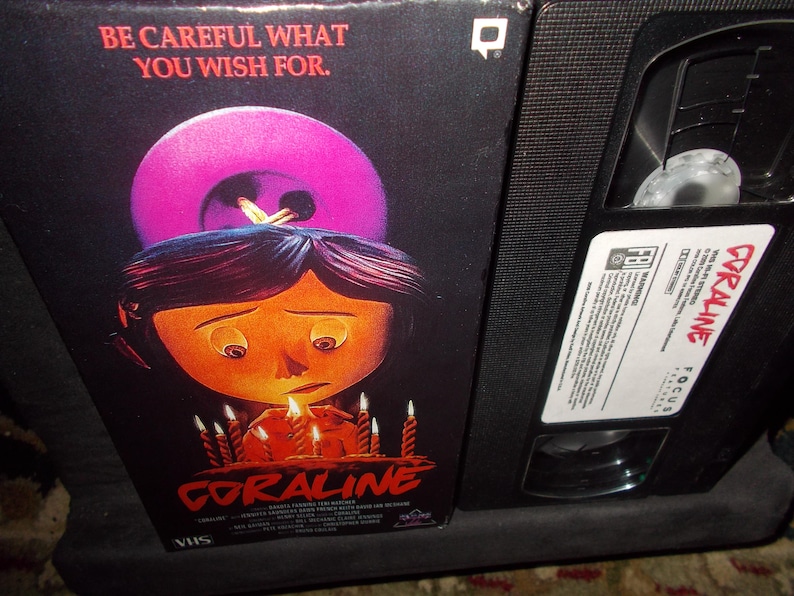 May include: A VHS tape and box set for the animated film "Coraline." The box art features a sad-looking girl with candles and a large button. The text "BE CAREFUL WHAT YOU WISH FOR" is at the top.