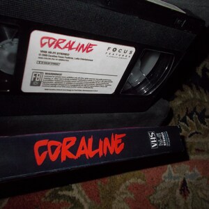 May include: A black VHS video cassette labelled "Coraline" in red text, with a matching case. The tape has a white label with additional text and a warning label. The case also has the movie title in red.