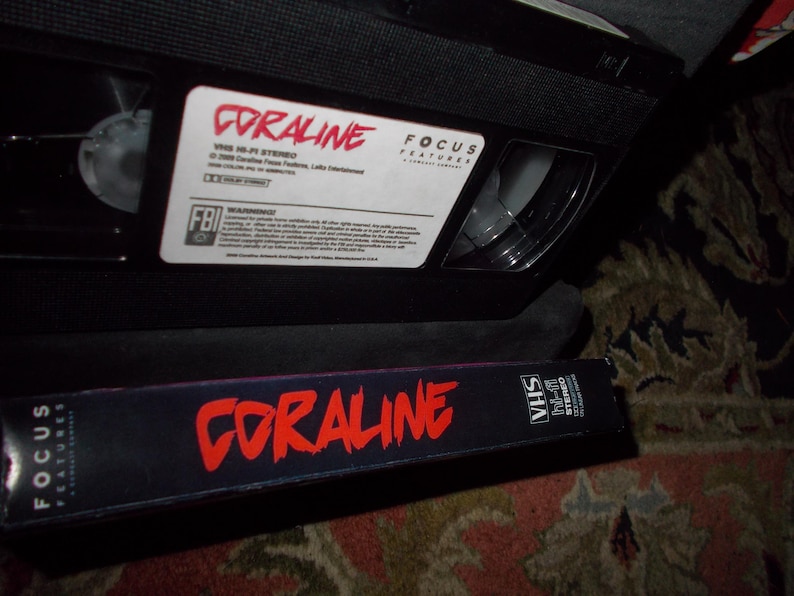 May include: A black VHS video cassette labelled "Coraline" in red text, with a matching case. The tape has a white label with additional text and a warning label. The case also has the movie title in red.