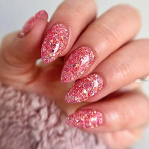 May include: Close-up of a hand with long, almond-shaped nails. The nails are painted with a vibrant pink glitter polish, featuring a mix of pink and gold glitter particles. The background is a soft, blurred pink.