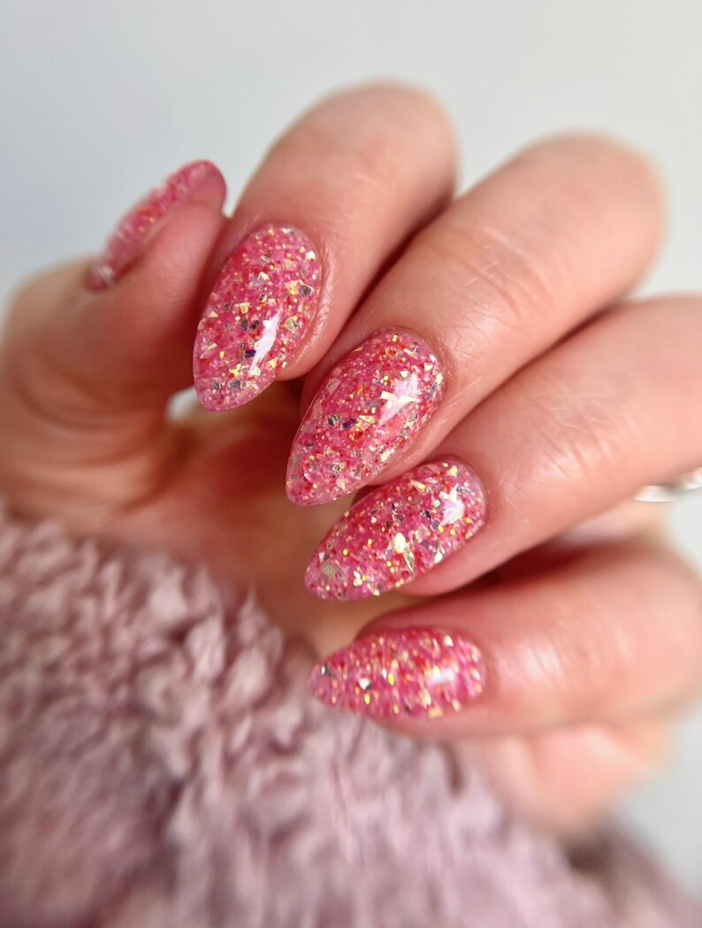 May include: Close-up of a hand with long, almond-shaped nails. The nails are painted with a vibrant pink glitter polish, featuring a mix of pink and gold glitter particles. The background is a soft, blurred pink.