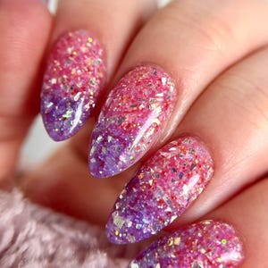 May include: Close-up of a hand with long, almond-shaped nails. The nails feature a gradient design, transitioning from a deep purple at the base to a vibrant pink at the tips. The nails are covered in sparkling glitter.