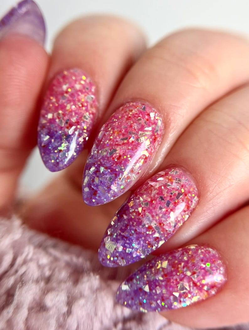 May include: Close-up of a hand with long, almond-shaped nails. The nails feature a gradient design, transitioning from a deep purple at the base to a vibrant pink at the tips. The nails are covered in sparkling glitter.
