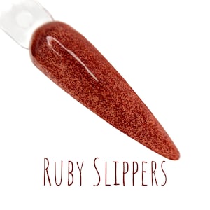 May include: A nail polish sample on a clear stick, displaying a deep red hue with a speckled finish. The words "RUBY SLIPPERS" are written in a dark red script below the sample, set against a white backdrop.