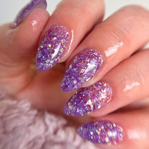 May include: Close-up of a hand with long, almond-shaped nails. The nails are painted with a vibrant purple glitter polish, reflecting light. The background is a soft, blurred pink.