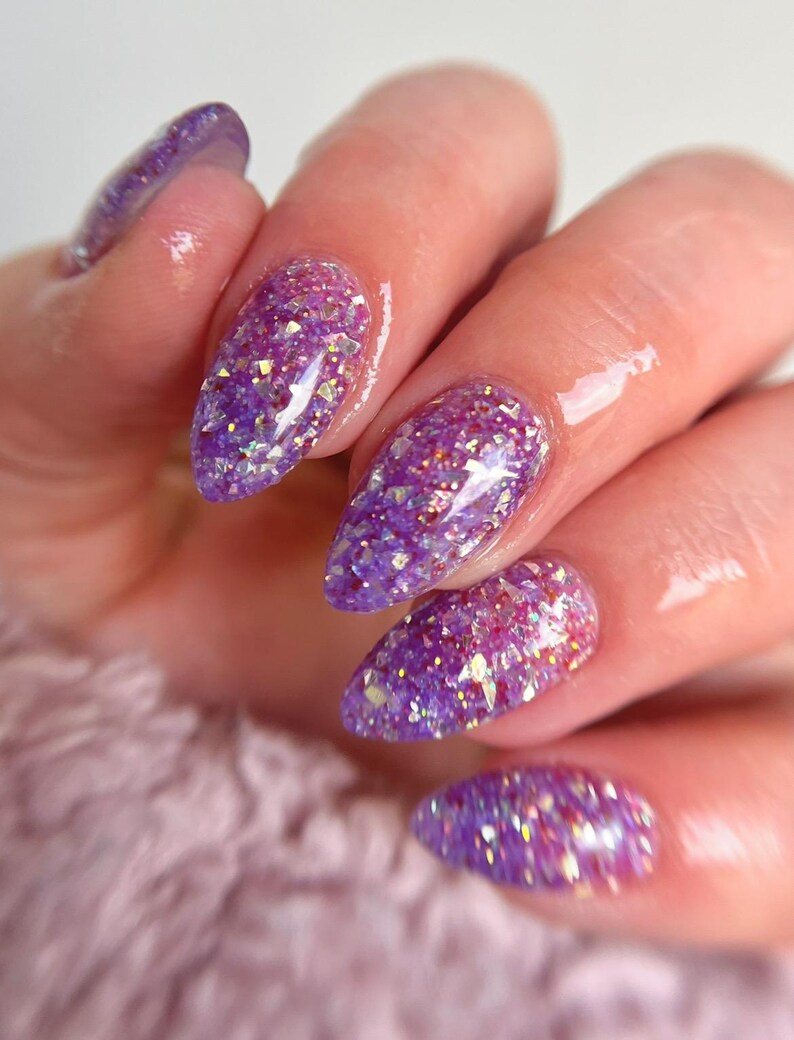 May include: Close-up of a hand with long, almond-shaped nails. The nails are painted with a vibrant purple glitter polish, reflecting light. The background is a soft, blurred pink.