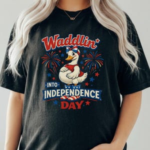 May include: Black t-shirt with a cartoon duck in patriotic attire, and the text "Waddlin' into Independence Day." The design incorporates fireworks, stars, and red, white, and blue colours, ideal for celebrating the Fourth of July.