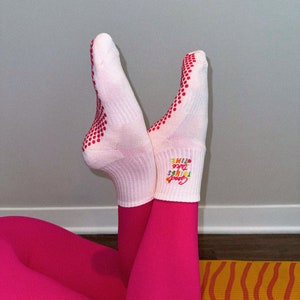 Pu&ograve; includere: Calzini bianchi con puntini rossi e il testo "Good Vibes Take Time" in colori arcobaleno. I calzini sono indossati da una persona che indossa leggings rosa e siede su un tappetino da yoga giallo e arancione.