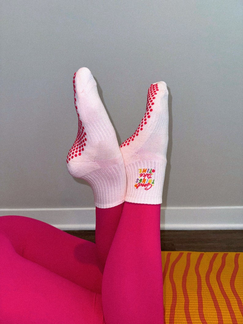 Pu&ograve; includere: Calzini bianchi con puntini rossi e il testo "Good Vibes Take Time" in colori arcobaleno. I calzini sono indossati da una persona che indossa leggings rosa e siede su un tappetino da yoga giallo e arancione.