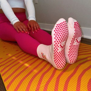 Pu&ograve; includere: Un paio di calzini bianchi con puntini antiscivolo rosa sul fondo. I calzini hanno un piccolo logo rosa che dice "SM" sul lato. I calzini sono su un tappetino da yoga giallo e arancione.