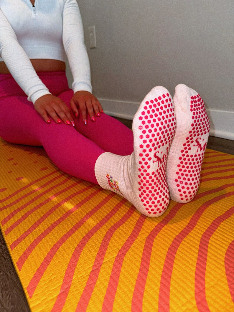 Pu&ograve; includere: Un paio di calzini bianchi con puntini antiscivolo rosa sul fondo. I calzini hanno un piccolo logo rosa che dice "SM" sul lato. I calzini sono su un tappetino da yoga giallo e arancione.