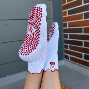 May include: White socks with red grippy dots on the bottom and a red heart design on the ankle. The socks are worn by a person with bare legs.
