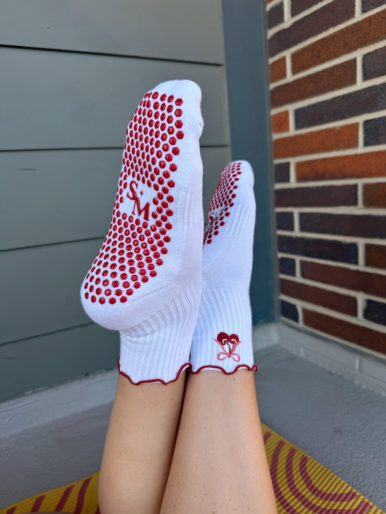 May include: White socks with red grippy dots on the bottom and a red heart design on the ankle. The socks are worn by a person with bare legs.