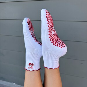 May include: White ankle socks with red grippy dots on the bottom and a red heart design on the ankle. The socks have a ruffled edge.
