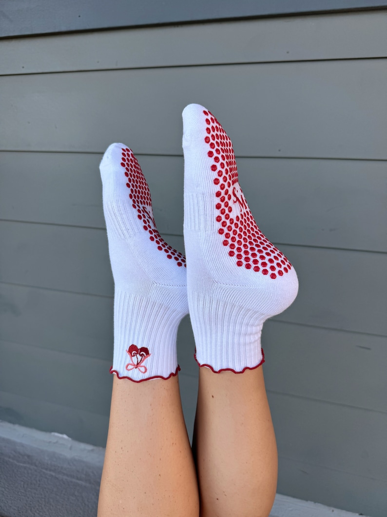 May include: White ankle socks with red grippy dots on the bottom and a red heart design on the ankle. The socks have a ruffled edge.