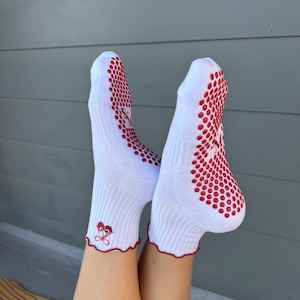 May include: White socks with red grippy dots on the bottom and a red embroidered heart design on the ankle. The socks are worn on a person's feet.