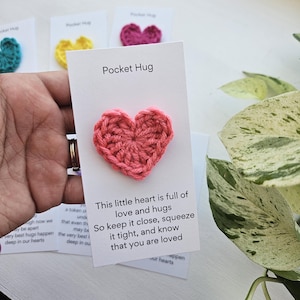 May include: A small crocheted pink heart attached to a white card with the text "Pocket Hug" and a message that reads "This little heart is full of love and hugs. So keep it close, squeeze it tight, and know that you are loved."