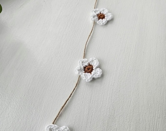 Puede incluir: Una guirnalda de flores blancas tejidas a crochet con centros marrones. La guirnalda está enhebrada en un cordel y cuelga sobre un fondo blanco.