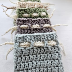 Puede incluir: Una pila de bolsas de ganchillo hechas a mano en varios colores, incluyendo verde, gris y morado. Cada bolsa presenta un patrón de puntada texturizada y un diseño crema contrastante. Las bolsas tienen extremos de hilo sueltos.