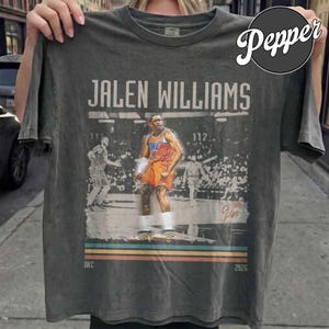 Op de afbeelding: Donkergrijs t-shirt met een afbeelding van basketballer Jalen Williams in een oranje en blauw tenue. Het shirt bevat de tekst "Jalen Williams", "OKC" en "2026", met een vintage ontwerp.