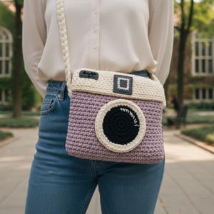 May include: A crochet camera-shaped crossbody bag in lavender, cream, and black. The bag features a large lens detail, a viewfinder, and a cream-colored strap. The bag is worn over the shoulder.