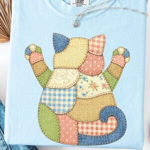 Op de afbeelding: Lichtblauw T-shirt met een patchwork kattenontwerp. De kat is gemaakt van verschillende stofpatronen, waaronder ruitjes, bloemen en stippen. Een zilveren ketting met een zonnehanger ligt op het shirt. Het shirt is gelabeld "CHAMBRAY".