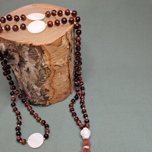 Może przedstawiać: Bransoletka z brązowych i czerwonych koralików z frędzlem i białymi muszelkami. Bransoletka jest prezentowana na drewnianym kłodzie.