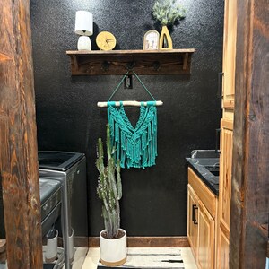 May include: A laundry room with a black textured wall, a wooden shelf with decor, and a macrame wall hanging. A tall cactus in a white pot sits on a black and white striped rug. Wooden cabinets and a washing machine are also visible.