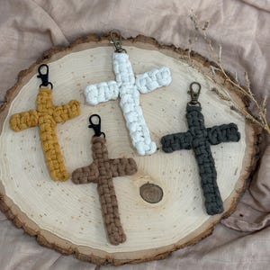 May include: Four macrame cross keychains in different colors: white, brown, mustard yellow, and dark green. The keychains are displayed on a wooden slice.