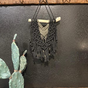 May include: A black macrame wall hanging with a natural wood branch and a central olive green design. The macrame features intricate knotting and fringe, creating a textured, bohemian aesthetic. It hangs against a dark, textured wall.