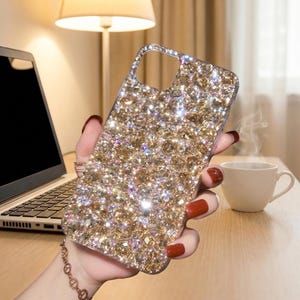 May include: A phone case covered in sparkling gold and clear crystals. The case has a rectangular shape with rounded corners and a cutout for the camera. The case is held in a person's hand.