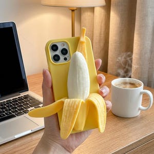 May include: A yellow phone case with a 3D banana design. The case features a peeled banana with a white fruit and yellow peel. The phone case is held in a hand, with a laptop and a cup of coffee in the background.