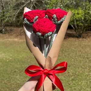 May include: A bouquet of three crocheted red roses with green leaves, wrapped in brown paper and tied with a red ribbon.