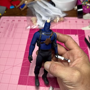 May include: A blue action figure with black trousers, boots, and gloves. The figure has a brown holster and a knife. The figure is missing a head and is being held by a person.