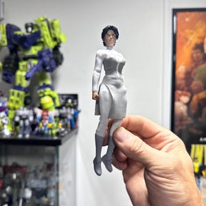 Op de afbeelding: Een grijs en wit actiefiguur van een vrouw in een futuristisch kostuum. Het figuur wordt in een hand gehouden, met de tekst "OMNI-CAVE" zichtbaar op de achtergrond. Andere actiefiguren zijn zichtbaar op de achtergrond.