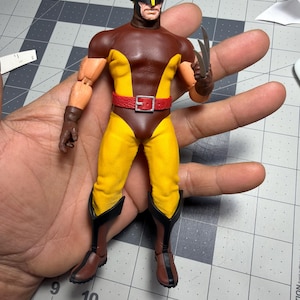 May include: A Wolverine action figure wearing a brown and yellow costume with a red belt, black mask, and brown gloves. The figure features three silver-colored claws extending from the left hand and stands against a cutting mat background.