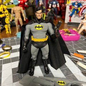 May include: A Batman action figure in a gray and black costume with a yellow bat symbol on the chest and belt. The figure is holding a gun and has a black cape. Additional costume pieces are visible in the foreground. Other action figures are in the background.