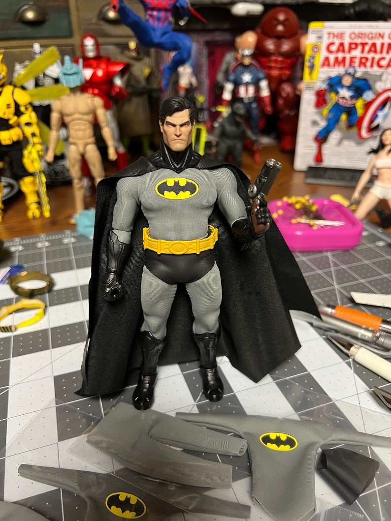 May include: A Batman action figure in a gray and black costume with a yellow bat symbol on the chest and belt. The figure is holding a gun and has a black cape. Additional costume pieces are visible in the foreground. Other action figures are in the background.