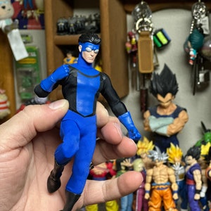 Puede incluir: Una figura de acción azul y negra de un superhéroe con una máscara azul y un traje azul y negro. La figura se sostiene en una mano y está rodeada de otras figuras de acción en un estante.
